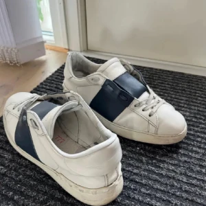 Valentino Garavani vita sneakers - Snygga vita sneakers från Valentino Garavani med bred marinblå skinnpanel över mitten. Klassisk rund tå, vita snören och diskret logga på plösen. Skorna har en stilren och modern look med exklusiv känsla.