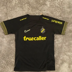 AIK svart matchtröja Nike M - AIK:s officiella svarta matchtröja från Nike med gula detaljer och klubbmärke på bröstet. Truecaller och SVEA som sponsors, samt gula mönster på ärmsluten. Tillverkad i lätt och ventilerande polyester, perfekt för fotboll eller att bära på läktaren.
