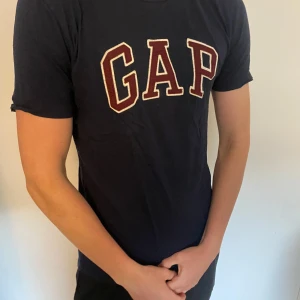 Mörkblå GAP t-shirt i bomull - Snygg mörkblå t-shirt från GAP med klassisk logga i vitt och vinrött framtill. Inte använd så är i princip ny. Mycket bra skick! 