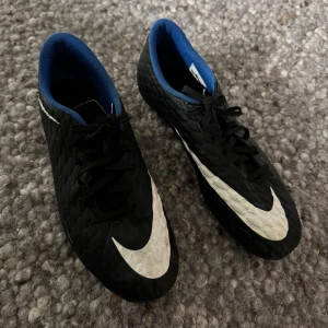 Nike Hypervenom svarta fotbollsskor - Säljer ett par Nike Hypervenom fotbollsskor i svart med stora vita swoosh-loggor och blå insida. Skorna har snörning och en texturerad ovandel i syntetmaterial för extra grepp på planen. Perfekta för dig som vill sticka ut på matchen! Skorna är perfekt skick, storlek 45