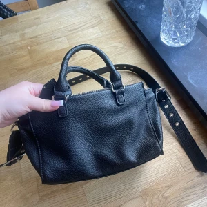 Svart handväska från Misako - Snygg svart handväska från Misako i strukturerat skinnimitation. Väskan har både handtag och justerbar axelrem, silverfärgade metalldetaljer och dragkedja upptill samt en extra ficka med dragkedja på baksidan. Perfekt storlek för vardagens essentials.