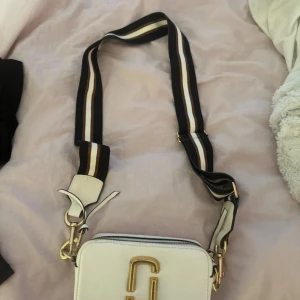 Vit axelväska Marc Jacobs med ränder - Säljer en snygg vit axelväska från Marc Jacobs med bred, randig axelrem i svart, brunt och vitt. Väskan har guldiga detaljer, dubbla dragkedjor och det klassiska Marc Jacobs-logot på både rem och väska. Perfekt för dig som vill ha en stilren men ändå cool accessoar. Original påse medföljer!