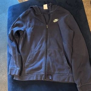 Mörkblå hoodiejacka från Nike XL - Mörkblå hoodiejacka från Nike i storlek XL, med dragkedja framtill och två stora fickor. Klassisk Nike-logga på bröstet och mjukt material som känns skönt mot huden. Perfekt för chill dagar eller när du vill ha en sportig look.