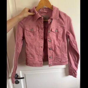 Rosa jeansjacka från Vero Moda XS - Snygg rosa jeansjacka från Vero Moda i storlek XS. Klassisk modell med knappar framtill, två bröstfickor med lock och långa ärmar. Jackan har en cool, croppad passform och är tillverkad i bomull. Perfekt för dig som vill sticka ut lite extra!