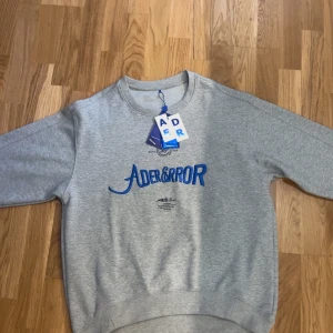 Grå sweatshirt från ADER ERROR - Snygg grå sweatshirt från ADER ERROR med blå broderad logga på bröstet. Klassisk rund halsringning och långa ärmar. Perfekt för dig som gillar streetwear och vill ha en stilren men ändå unik look. Helt oanvänd och lapparna kvar. Kostar över 3000 kr ny 