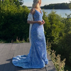 Blå glittrig långklänning med v-ringning - Magisk blå långklänning med glittriga paljetter över hela tyget. Klänningen har smala axelband, v-ringning och figurnära passform som faller snyggt mot kroppen. Perfekt för bal eller fest där du vill glänsa extra mycket.