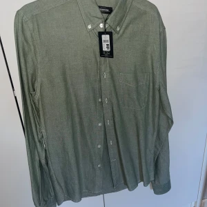 Grön oxfordskjorta från Dressmann - Snygg grön oxfordskjorta från Dressmann i slim fit-modell. Skjortan har klassisk button down-krage, bröstficka och vita knappar framtill. Tillverkad i bomull och passar perfekt till jeans eller chinos.