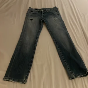Blå bootcut jeans från Lee - Snygga blå jeans från Lee med bootcut passform och slitna detaljer. Jeansen har klassisk femficksdesign, dragkedja och knapp framtill samt Lee-lapp bak i midjan. Tillverkade i jeansmaterial med en tvättad look och ljusare partier.