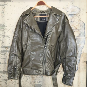Skinnjacka från Zara - Snygg olivgrön skinnjacka från Zara med klassisk bikerstil, breda slag, dragkedjor på ärmarna och bälte i midjan. Jackan har en cool oversized passform och detaljerade sömmar som ger extra edge till din look.