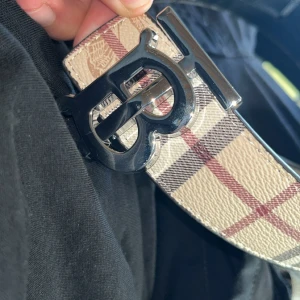Burberry bälte - Snyggt Burberry-bälte med klassiskt rutigt mönster i beige, svart och rött. Bältet har en stor, blank silverspänne med TB-logga och är gjort i skinn. Perfekt för att lyfta vilken outfit som helst med en lyxig touch.