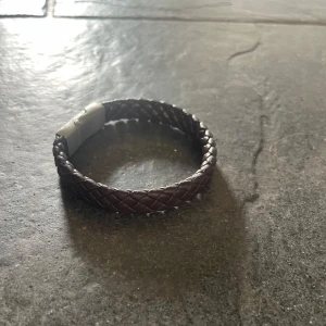 Brunt flätat läderarmband - Snyggt armband i mörkbrunt flätat läder med ett stilrent metallspänne. Perfekt accessoar för en cool och avslappnad look. Passar både tjejer och killar som gillar enkel men trendig stil.