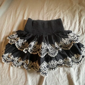 Svart volangkjol med broderad spets - Supersöt svart kortkjol med bred resår i midjan och flera lager volanger. Kjolen har vit broderad spets längs kanterna som ger en romantisk och lekfull vibe. Perfekt för dig som gillar detaljer och vill sticka ut lite extra.