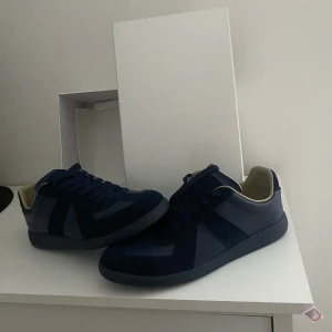 Mörkblå sneakers från Maison Margiela - Snygga mörkblå sneakers från Maison Margiela med klassisk siluett. Skorna har ovandel i skinn och mocka, mörkblå snörning och beige innerfoder. Sulan är i gummi med Maison Margiela-logga undertill. Perfekta för dig som gillar stilrena och exklusiva sneakers.