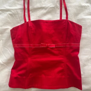 Röd korsett-topp med rosett - Supersnygg röd korsett-topp med smala axelband och markerad midja. Toppen har en söt liten rosett framtill och dekorativ söm längs midjan.