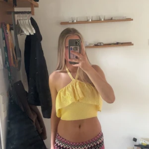 Gul croppad stickad topp med volang - Söt gul croppad stickad topp med halterneck och volangdetalj framtill. Toppen är ärmlös och har en luftig känsla, perfekt för varma dagar. Den har en smockad nederdel som sitter snyggt på kroppen och ger en trendig look.