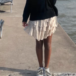 Beige volangkjol med blommigt mönster - Supersöt kort kjol i beige med småblommigt mönster och flera lager volanger. Kjolen har en flowy och lätt känsla, perfekt för varmare dagar. Den är feminin och trendig, passar dig som gillar romantisk stil.