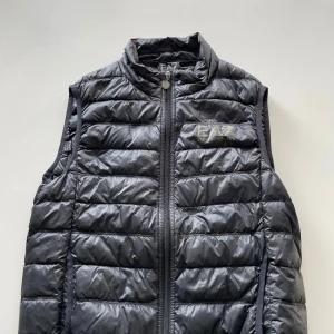 Svart dunväst från EA7 Emporio Armani - Snygg svart dunväst från EA7 Emporio Armani med quiltad design och hög krage. Västen har dragkedja framtill och diskret logga på bröstet. Tillverkad i polyester och dun, perfekt för lager-på-lager-stil under kyliga dagar.
