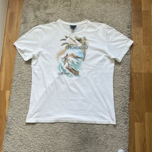Vit t-shirt Ralph Lauren Honolulu print - Vit t-shirt från Ralph Lauren med färgglatt tryck på bröstet som visar en surfare, palmer och texten 'A Trip to Honolulu'. Sitter mer som M men kan passa LKlassisk passform med rund hals och korta ärmar. Mjuk bomullskvalitet som känns skön mot huden. Priset är inte hugget i sten!