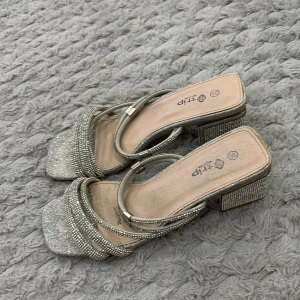 Glittriga silver sandaler med klack - Snygga sandaler i silver med glittriga remmar och fyrkantig tå. Skorna har blockklack täckt av små stenar för extra bling och flera tunna remmar med strassdetaljer. Perfekta för att sticka ut och ge din outfit en extra touch.