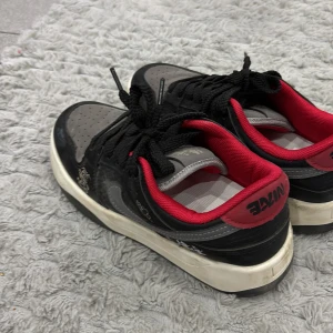 Nike sneakers svart/grå med röd detalj - Snygga Nike sneakers i svart och grått med röda detaljer på häl och insida. Skorna har klassisk snörning, vit sula och grå swoosh på sidan. Ovandelen är i mocka och syntet med perforerad tåbox för extra ventilation. Perfekta för dig som gillar streetstyle.