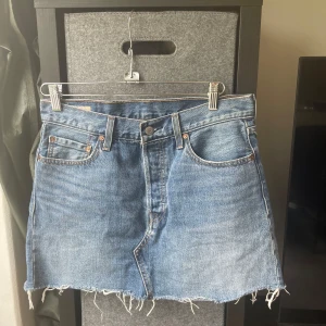 Levi's jeanskjol äkta - Snygg blå jeanskjol från Levi's med klassiska fem fickor och rå, fransig kant nertill. Kjolen har bälteshällor, knapp och dragkedja framtill samt Levi's ikoniska läderpatch bak. Storlek 28