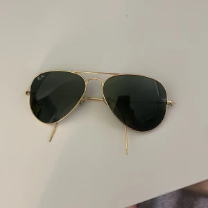 Ray-Ban Aviator guld solglasögon - Snygga Ray-Ban Aviator solglasögon med klassisk pilotform. Tunn guldfärgad metallbåge och mörkgröna glas. Modellnummer RB 6049, storlek 55 mm. Perfekta för dig som vill ha en tidlös och cool look.