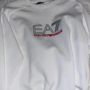 Vit sweatshirt från Emporio Armani - Snygg vit sweatshirt från Emporio Armani med EA7-logga i svart och rött på bröstet. Perfekt för en stilren och avslappnad look.
