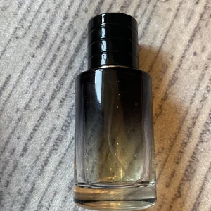 Svart parfymflaska med rund form - Stilren parfymflaska i glas med svart, rund kork och mörktonad design som går från svart upptill till transparent nedtill. Flaskan har en modern look och passar perfekt på hyllan för dig som gillar minimalistisk stil.