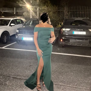 Mörkgrön offshoulder långklänning med slits - Säljer en elegant mörkgrön långklänning med offshoulder-modell och hög slits framtill. Klänningen har figurnära passform och är perfekt för bal eller fest. Stilren design utan mönster och med en snygg axelbandslös look.