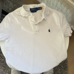 Vit pikétröja från Ralph Lauren - Säljer en klassisk vit pikétröja från Ralph Lauren i slim fit. Tröjan har en broderad logga på bröstet och knappar vid kragen. Perfekt för en stilren look. 