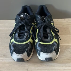 Adidas sneakers med gröna detaljer - Snygga Adidas sneakers i svart med gröna och grå detaljer. Skorna har en sportig design med snörning och en robust sula. Perfekta för en aktiv livsstil.