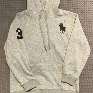 Polo Ralph Lauren Hoodie (Big Pony) - Säljer denna stilrena och eftertraktade grå hoodie från Ralph Lauren med en stor logga (Big Pony) på bröstet och siffran 3 på ärmen. Det är en gammal modell som inte säljs längre men den är knappt använd, max 3 gånger. Jättefint skick! Stl M men ganska slim fit så passar Stl S också. Kom gärna med frågor:) 