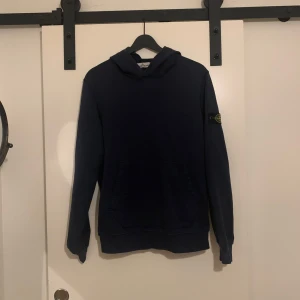 Mörkblå hoodie från Stone Island - Säljer en mörkblå hoodie från Stone Island med klassisk patch på vänster ärm. Tröjan har huva och magficka, perfekt för en avslappnad stil. Passar dig som gillar streetwear och vill ha något stilrent och bekvämt. Storlek 170 junior