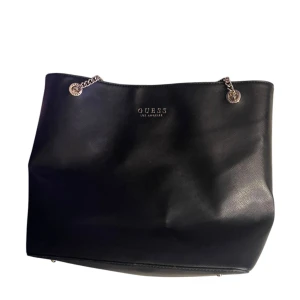 Svart axelväska från Guess - Snygg svart axelväska från Guess med kedjedetaljer och logotyp framtill. Väskan har en dragkedja och flera fack inuti för enkel organisering. Mycket bra skick, nästan ny då jag använt den 3-4 gånger, orginal pris ca 1200 kr