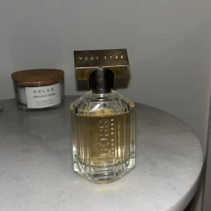 Hugo Boss The Scent Eau de Parfum - En elegant parfym från Hugo Boss, The Scent. Flaskan är transparent med guldtonad vätska och har en metallisk, rektangulär kork. Designen är stilren och lyxig med tydlig logotyp på framsidan.