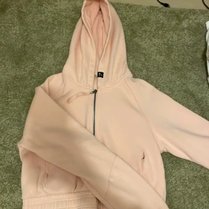 Ljusrosa hoodie med dragkedja - Säljer en ljusrosa hoodie med huva och dragkedja framtill. Tröjan har långa ärmar och en mjuk, skön känsla. Perfekt för en avslappnad stil och enkel att matcha med jeans eller leggings.