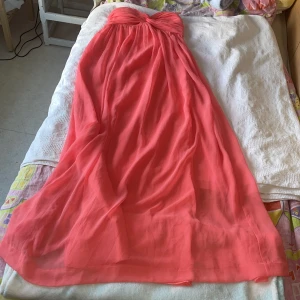 Korallfärgad axelbandslös klänning från Nelly Trend - Säljer en vacker korallfärgad axelbandslös klänning från Nelly Trend. Klänningen har en elegant twistdetalj vid bysten och ett flödande långt fall. Perfekt för sommarens festligheter!