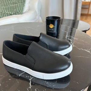 Svarta slip-on sneakers med vit sula - Säljer ett par svarta slip-on sneakers med vit, platt sula. Skorna har en stilren och enkel design utan snörning, med elastiska partier på sidorna för enkel påtagning. Perfekta för en clean och avslappnad look. Aldrig använda. Jag har bara för många skor😅 storlek 39.