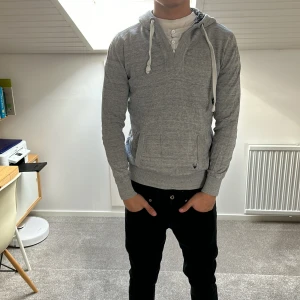 Grå hoodie med dragkedja och snörning - Säljer en stilren grå hoodie med huva och vita snören. Tröjan har dragkedja framtill, fickor på sidorna och ribbade muddar vid ärmslut och nederkant. Perfekt för en avslappnad look.