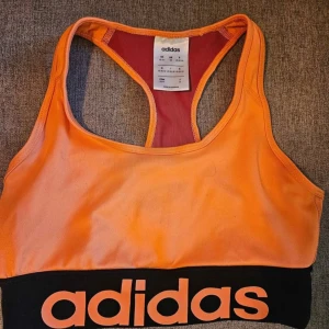 Orange sport-bh från Adidas - Säljer en sportig orange sport-bh från Adidas med bred svart resår och stor logga framtill. Brottarrygg och kontrastfärgad insida i rött. Perfekt för träning och ger bra stöd.
