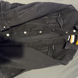 Svart jeansjacka från Calvin Klein - Snygg svart jeansjacka från Calvin Klein med klassiska knappar framtill. Jackan har en stilren design med långa ärmar och är perfekt för en cool och avslappnad look. Märkesdetaljer syns på etiketten och insidan av jackan.