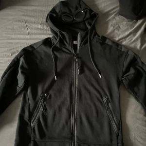 Svart hoodie med dragkedja och glasögon - Snygg svart hoodie med dragkedja och inbyggda glasögon i huvan. Perfekt för en unik stil. Den har långa ärmar och praktiska fickor med dragkedja. Passar bra för både vardag och speciella tillfällen.