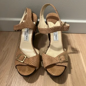 Beige sandaler med klack från Jimmy Choo - Säljer ett par snygga beige sandaler från Jimmy Choo med hög klack och platåsula. Skorna har ormskinnsmönster, guldfärgade nitar och spännen samt öppen tå. Perfekta för att lyfta din outfit! Dem är i storlek 40 och köpta för 6000 men säljer för 1400
