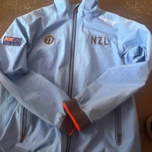 Ljusblå vindjacka NZL med dragkedja. Sail racing - Säljer en ljusblå vindjacka från NZL med hel dragkedja, höga muddar och detaljer i mörkgrått. Jackan har broderade märken, två fickor med dragkedja och en patch med flagga på ärmen. Perfekt för dig som gillar sportig stil.