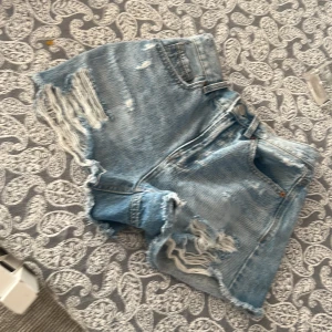 Blå jeansshorts med slitningar - Säljer ett par coola blå jeansshorts med slitningar och fransiga kanter. Perfekta för en avslappnad sommarlook. De har en klassisk femficksdesign och knappgylf.