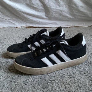 Svarta och vita sneakers från Adidas - Snygga svarta sneakers från Adidas med de klassiska tre vita ränderna på sidan. Skorna har en vit sula och snörning framtill. Perfekta för en avslappnad stil.
