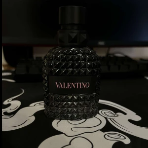 Svart parfymflaska från Valentino - Elegant svart parfymflaska från Valentino med ett unikt, nitat mönster som ger en lyxig känsla. Flaskan har en stilren design med en svart etikett och text i rosa. Perfekt för den som vill ha en sofistikerad doftupplevelse.