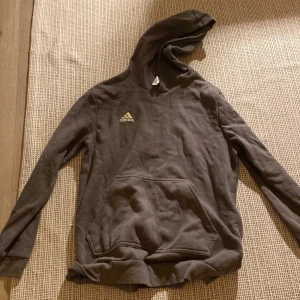 Olivgrön hoodie från Adidas - Mysig grå hoodie från Adidas med klassisk logga på bröstet. Tröjan har huva, magficka och långa ärmar. Perfekt för en avslappnad stil och enkel att matcha med andra plagg.