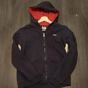 Mörkblå hoodie från FILA med dragkedja - Säljer en mörkblå hoodie från FILA med vit och röd logga på ryggen och liten logga fram. Tröjan har dragkedja, huva med röd insida och fickor framtill. Använd fåtal gånger, skulle klassa storleken som en xs/s