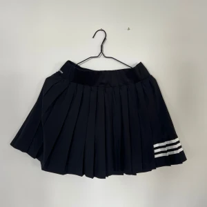 Svart plisserad tenniskjol från Adidas - Säljer en svart plisserad padelkjol från Adidas med inbyggda shorts. Kjolen har tre vita ränder på ena sidan och Adidas-logga framtill. Perfekt för dig som gillar sportig stil och vill ha något bekvämt och snyggt.